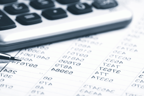 A black and white picture of the corner of a calculator and the tip of a pen on a piece of paper with multiple numbers in list format.