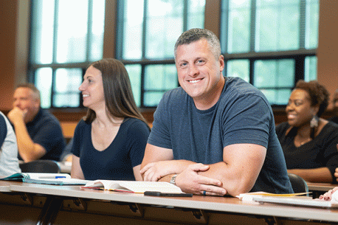 A middle-aged white male with short greying hair and a grey t-shirt sitting at a table in a classroom looks toward the camera and smiles with a diverse group of classmates in the background.  