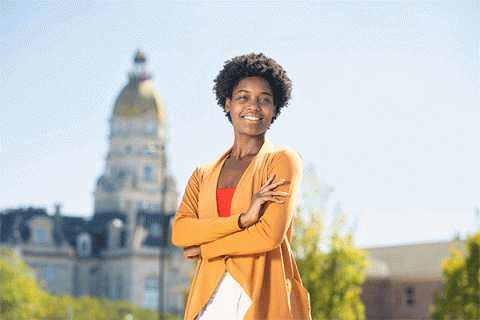 A smiling, tall Black female with short, dark, curly hair stands outside on a sunny day with the Vigo County Courthouse visible but slightly out of focus behind her. She has her arms crossed and wears a long, burnt-orange jacket over a red t-shirt and a white skirt. Trees and greenery are also visible around the courthouse and other buildings behind her.  